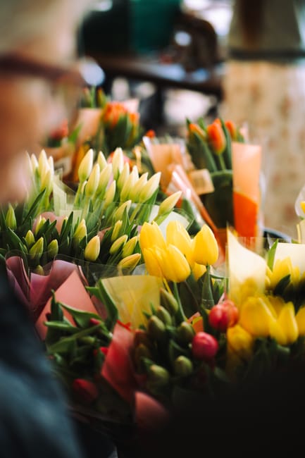 A vibrant display of fresh flowers including yellow tulips, white tulips, and pink buds arranged together in bouquets. The flowers are wrapped in pastel-colored tissue paper and clear cellophane, with