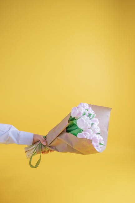 A person's hand, dressed in a white shirt, holds a bouquet of fresh tulips wrapped in light brown tissue paper with a green ribbon against a solid yellow background. The tulips are light pink with hin