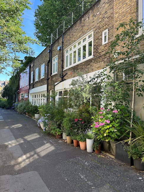 A row of potted flowering plants and lush greenery arranged along the sidewalk outside a residential building with large white-framed windows and a brick facade. The plants include pink hydrangeas, wh