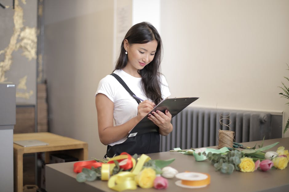 A professional florist with long dark hair, wearing a white t-shirt and black apron, is writing on a clipboard while standing at a table with various fresh flowers, including yellow roses, pink tulips