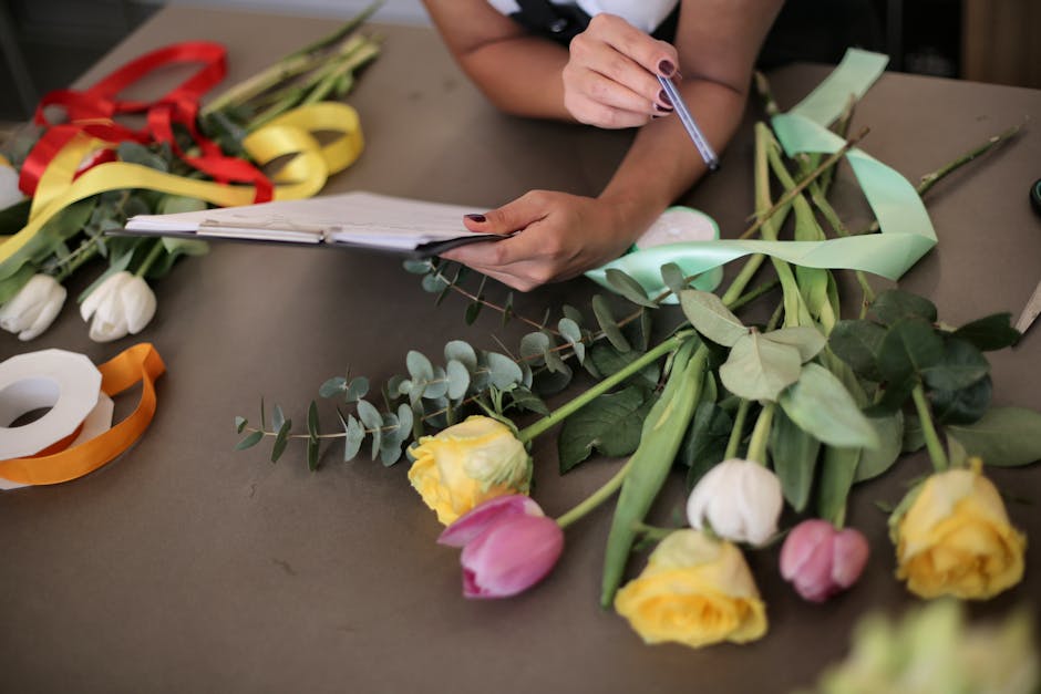 Close-up of a florist's workspace featuring a variety of fresh flowers including yellow and pink tulips, surrounded by green foliage and wrapped ribbons, with a person holding a pen and referencing a