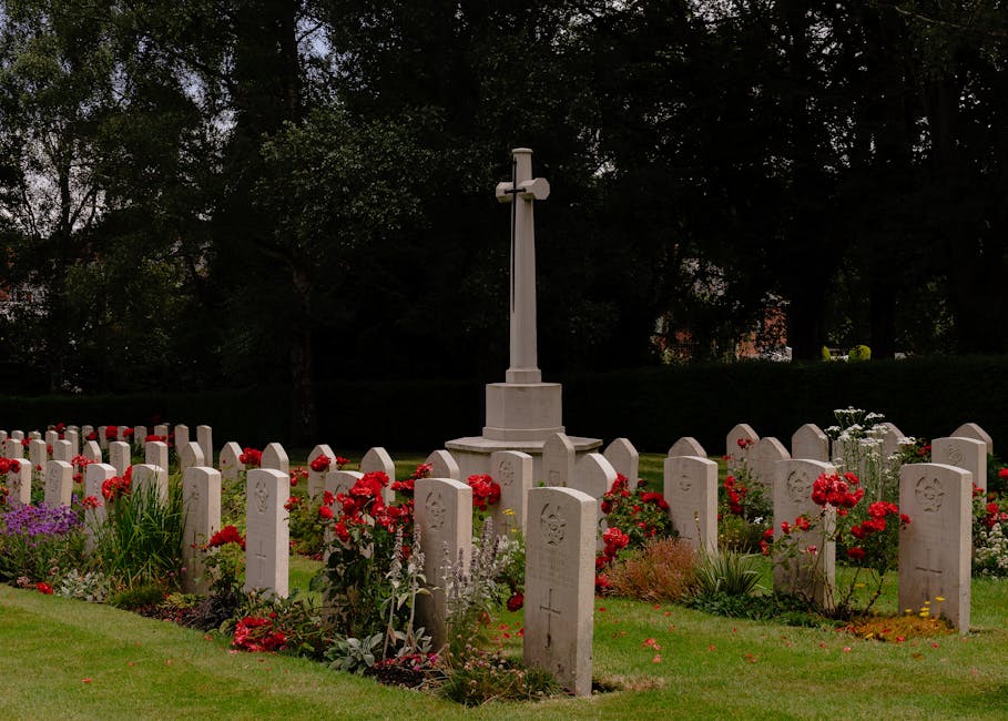 A memorial site featuring multiple white gravestones arranged in neat rows, surrounded by vibrant red and purple flowers. At the center stands a tall white cross monument, set against a backdrop of ta