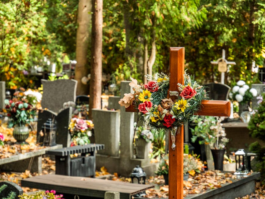 A wooden cross decorated with a floral arrangement sits amidst a graveyard surrounded by various headstones and monuments. The bouquet features red roses, yellow daisies, and soft pink and cream flowe