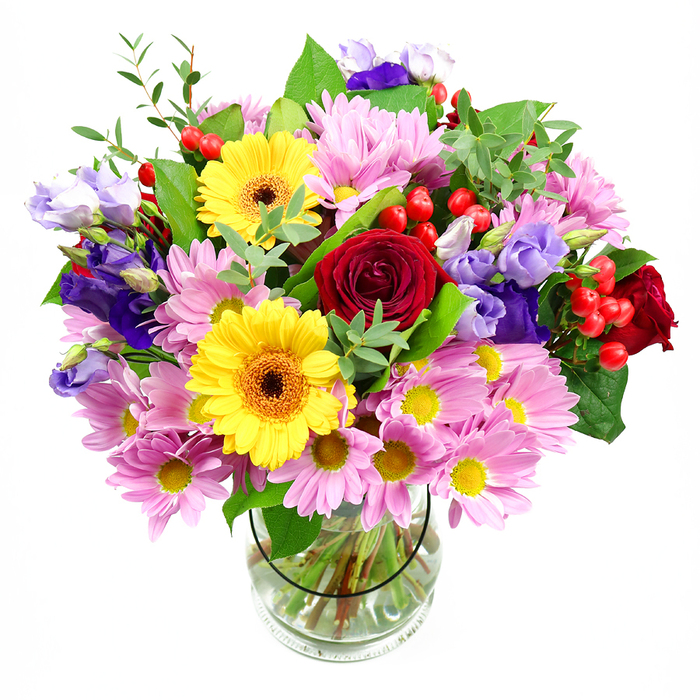 In this image, a lush, dome-shaped bouquet bursts from a clear glass vase, set crisply against a white background so every nuance of colour and texture is visible. The focal point of the design is a cluster of deep red roses, their velvety petals spiralling inward to create a sense of rich romance and classic elegance. Tucked just beside them, two bright yellow gerbera daisies with dark brown-orange centres seem to smile outward, infusing the entire piece with cheer, like sunshine streaming across a quiet Golders Green garden on a clear morning. Surrounding these central flowers, generous sprays of soft pink daisies or chrysanthemums form a halo of pastel colour; each bloom is made of numerous slender petals, cupping a small yellow centre that echoes the gerberas and adds a subtle, harmonious glow. Interlaced among the daisies and roses are graceful purple and lavender lisianthus, their gently fluted, bell-shaped flowers opening at different stages, creating depth and a slightly whimsical rhythm. Bright red hypericum berries appear here and there in rounded clusters, their glossy skins offering a striking, textural contrast to the softness of the petals. Various types of foliage frame and support the blooms: deep green, broad leaves give the design structure, while lighter, more delicate stems lace through the bouquet, adding airiness and a freshly picked feel. Through the transparent vase, an orderly grid of stems rests in clear water, revealing the care taken by an experienced florist. The overall impression is of a joyful, thoughtfully balanced arrangement, perfect for marking birthdays, thank-you gestures, or celebrations at home near Brent Cross, bringing a burst of garden brightness indoors.